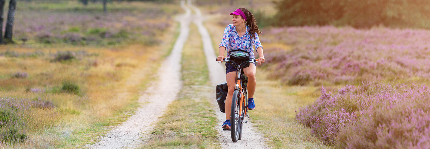 Fot van een sportieve jonge dame op de fiets over een bloeiend heideveld in Twente op een half verhard pad.